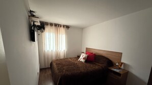 A bedroom with a bed, a nightstand, a lamp, a window with curtains, and a wall-mounted shelf.