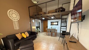 A modern living room with a black sofa, a dining table with chairs, a TV mounted on the wall, and a loft area above.