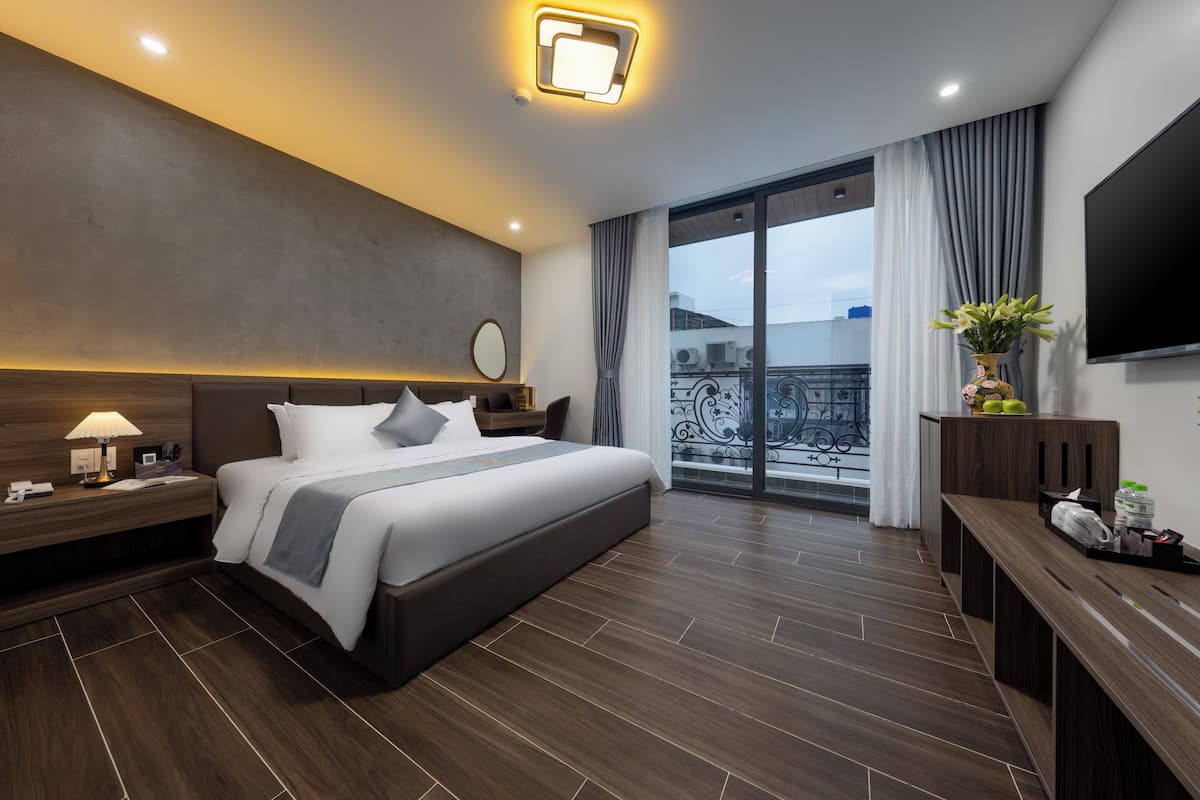A modern hotel room with a large bed, a bedside table with a lamp, a flat-screen TV mounted on the wall, and a balcony with a view.