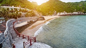 A coastal hotel with a stone pathway leading to the beach, surrounded by lush greenery and a mountainous backdrop. - Cat Ba Wistaria Hotel - Pool & Massage (Hai Phong)