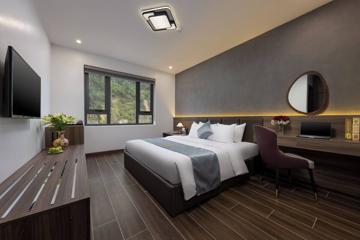 A modern hotel room with a large bed, a desk with a laptop, a flat-screen TV, a large window, and a mirror on the wall.