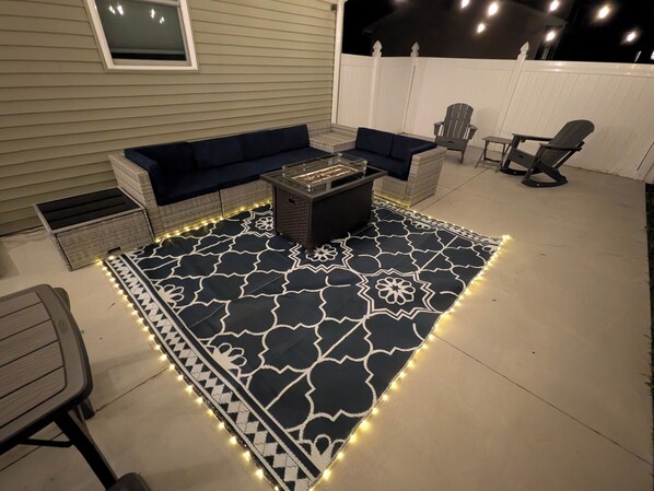 Un patio con un tapete estampado, muebles de exterior e iluminacion con luces colgantes.