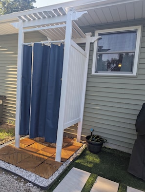 Ein überdachter Patio-Bereich mit Holzdeck, einer weißen Pergola und einem Fenster mit Blick auf Bäume.