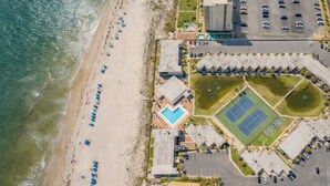 An aerial view of a coastal property with a beach, a swimming pool, tennis courts, and a parking lot.