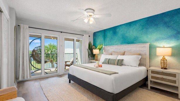 A bedroom with a large bed, a ceiling fan, a nightstand with a lamp, and a view of a balcony with outdoor furniture.