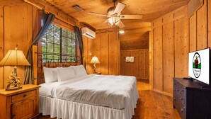 Un dormitorio estilo cabaña de madera con una cama, dos lámparas de noche, un ventilador de techo y un televisor sobre un aparador.