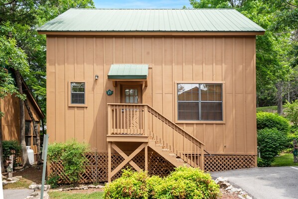 A tan wooden building with a green roof, a small porch, and a staircase leading to the entrance.