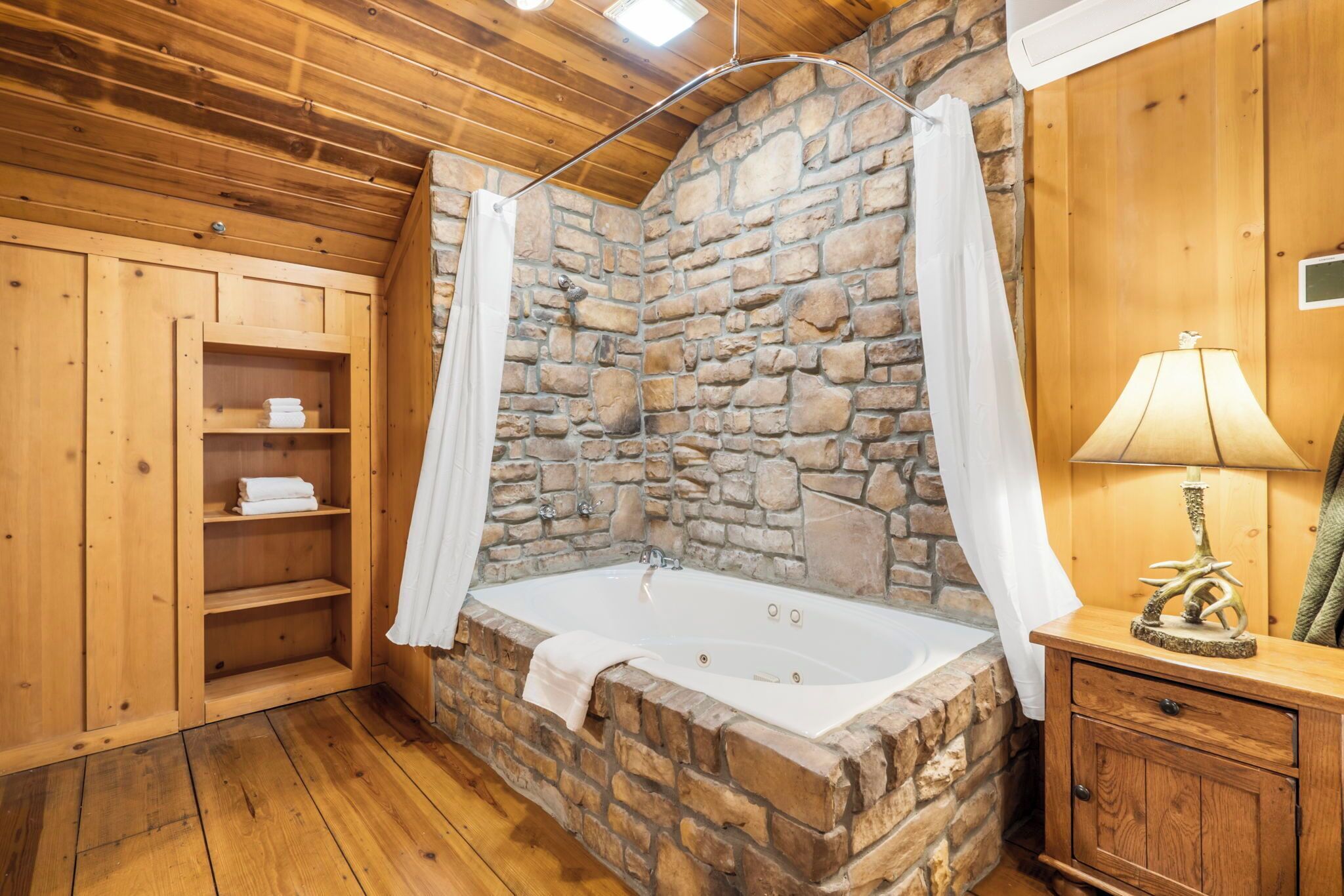 A stone-walled bathroom with a wooden floor, a bathtub, a wooden cabinet with shelves, and a lamp.