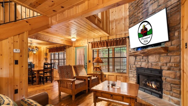 Un acogedor interior de cabaña con chimenea de piedra, muebles de madera y un televisor montado en la pared.