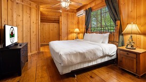 Un dormitorio de cabaña de madera con cama, mesitas de noche, ventilador de techo y ventana con cortinas.