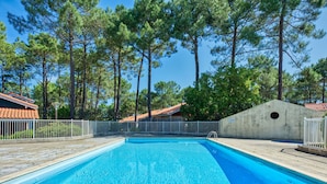 Ein rechteckiger Pool mit klarem, blauem Wasser, umgeben von einem weißen Zaun, vor einer Kulisse aus hohen Kiefern und einem Gebäude mit roten Ziegeln.