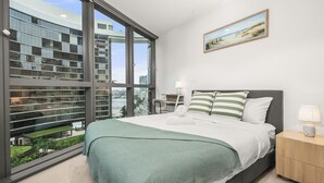 A modern bedroom with a large window, a bed with green and white bedding, a nightstand with a lamp, and a painting on the wall.