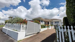 Une maison à deux étages avec une clôture en bois peinte en blanc, une véranda et une allée menant à l’entrée.