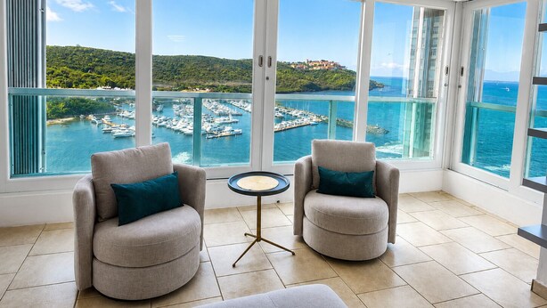 A balcony with two armchairs and a small table, overlooking a marina with boats and a hilly landscape. - Caribbean Dream Condo – Oceanfront, Mountain Views & Modern Comfort (Fajardo)
