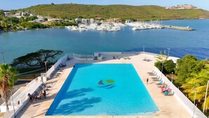 A large rectangular pool with a clear blue center, surrounded by a white deck with lounge chairs. In the background, there is a marina with boats and a hilly landscape. - Caribbean Dream Condo – Oceanfront, Mountain Views & Modern Comfort (Fajardo)