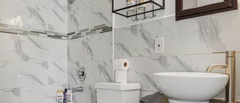 A bathroom with a marble shower wall, a freestanding bathtub, a white toilet, a vanity with a sink, and a mirror.