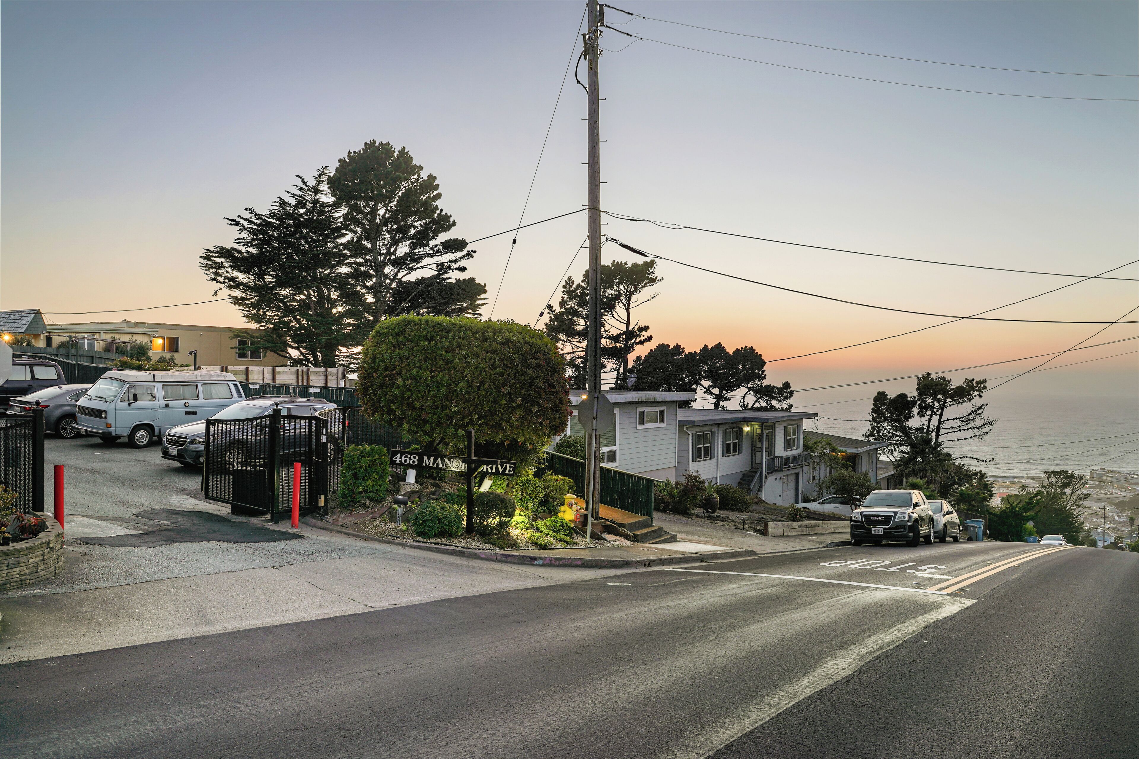 Una calle residencial con coches aparcados, un acceso con puerta de reja y una casa con vistas al océano.