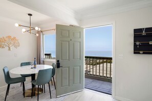 A dining area with a round table, teal chairs, and a light-colored floor. A door leads to a balcony with a view of the ocean. - Delightful 2BR Mountain OceanSunset View (Pacifica)