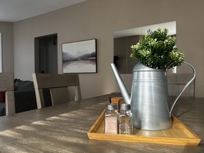 Ein Metall-Gießkännchen mit einer Pflanze auf einer Holzschale, zwei Salzstreuer und ein Bild an der Wand.