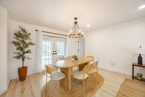 A dining area with a round wooden table, six chairs, a potted plant, a chandelier, and a door leading outside.