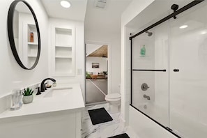 A modern bathroom with a white vanity, a black sink, a mirror, a toilet, and a glass shower enclosure.