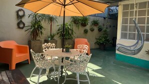 Une terrasse extérieure avec une table ronde et des chaises, entourée de plantes en pot et agrémentée d’un fauteuil suspendu.