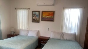 Une chambre avec deux lits, chacun recouvert d’une courtepointe bleu clair, avec des oreillers blancs et une petite table de chevet en bois sur laquelle se trouve une lampe. Deux tableaux encadrés ornent le mur : l’un représente un paysage tropical et l’autre, un coucher de soleil.