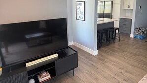 A modern living room with a flat-screen TV, a coffee table with books, and a wall-mounted sign that reads "MEET ME AT THE LAKE".