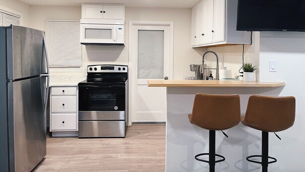 A modern kitchen with stainless steel appliances, a microwave, oven, and a built-in TV. There are two bar stools and a small bar area.