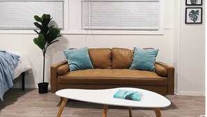 A living room with a brown leather sofa, two blue cushions, a white coffee table with a book, a potted plant, and a bed with a blue blanket.