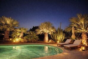 A nighttime pool area with palm trees, lounge chairs, and a clear pool.