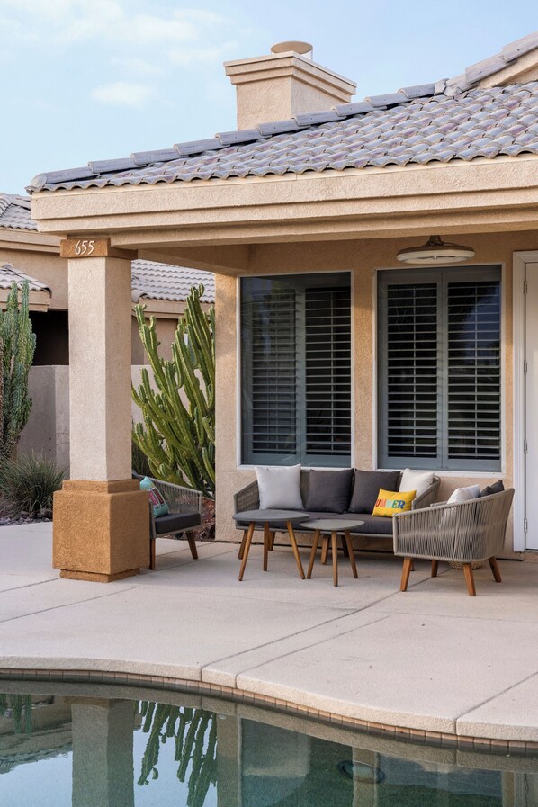 A covered outdoor seating area with a pool, a cactus, and a building with a tiled roof.