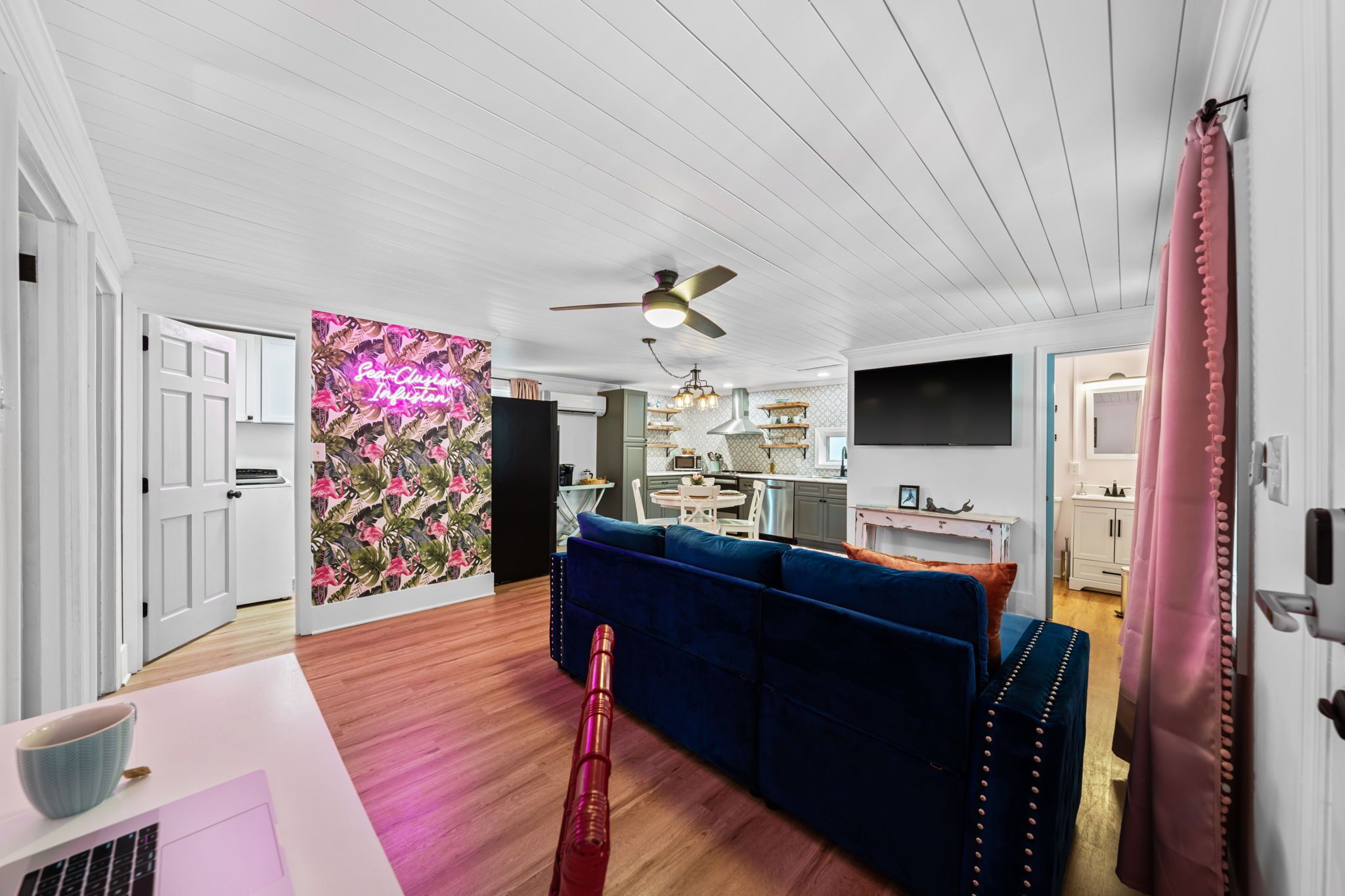 Un salon moderne avec un canapé bleu, un plancher en bois, un ventilateur de plafond et une cuisine en arrière-plan.
