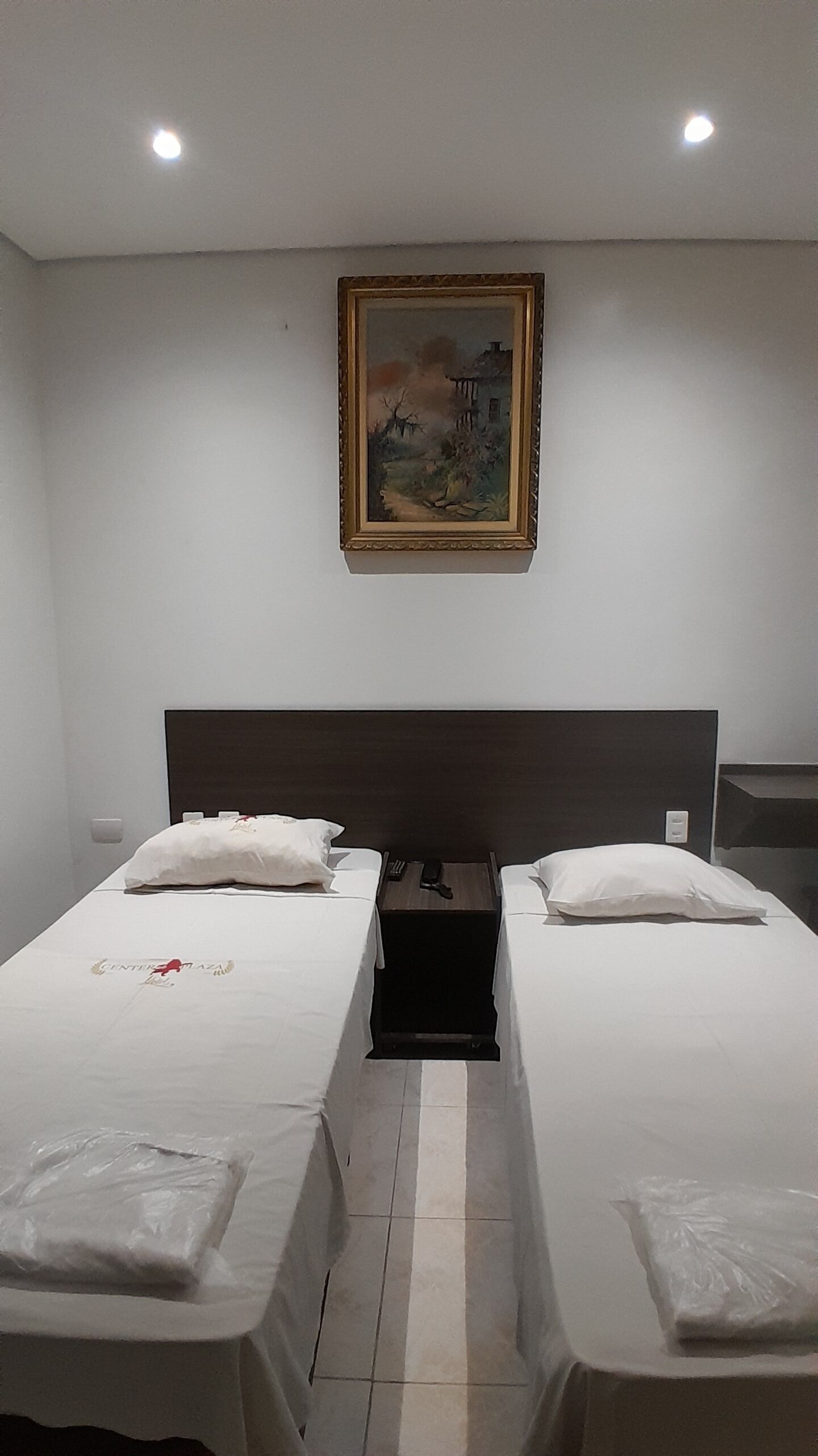 A hotel room with two single beds, a wooden headboard, a painting on the wall, and a nightstand with a lamp.