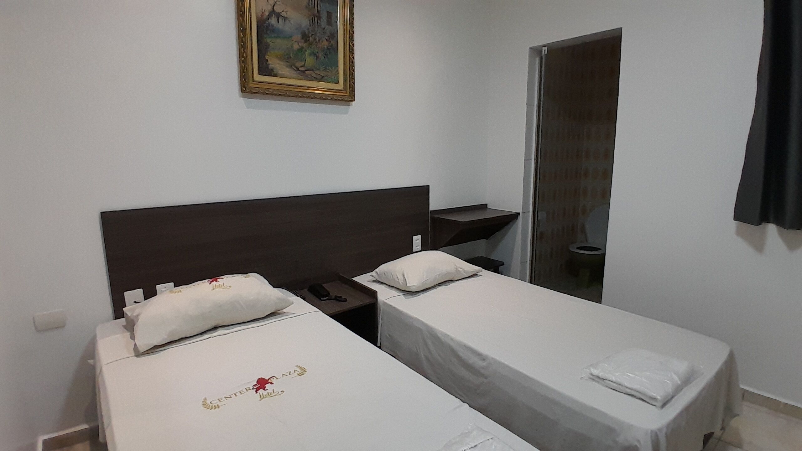 A hotel room with two single beds, a nightstand, a small table, a painting on the wall, and a bathroom door.