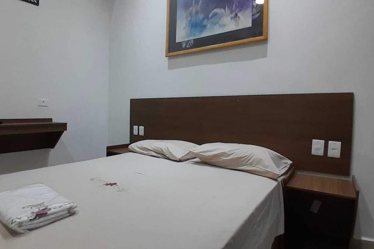 Una cama bien hecha con una colcha blanca y dos almohadas, un cabecero de madera, una mesita de noche con una lámpara y un cuadro en la pared.