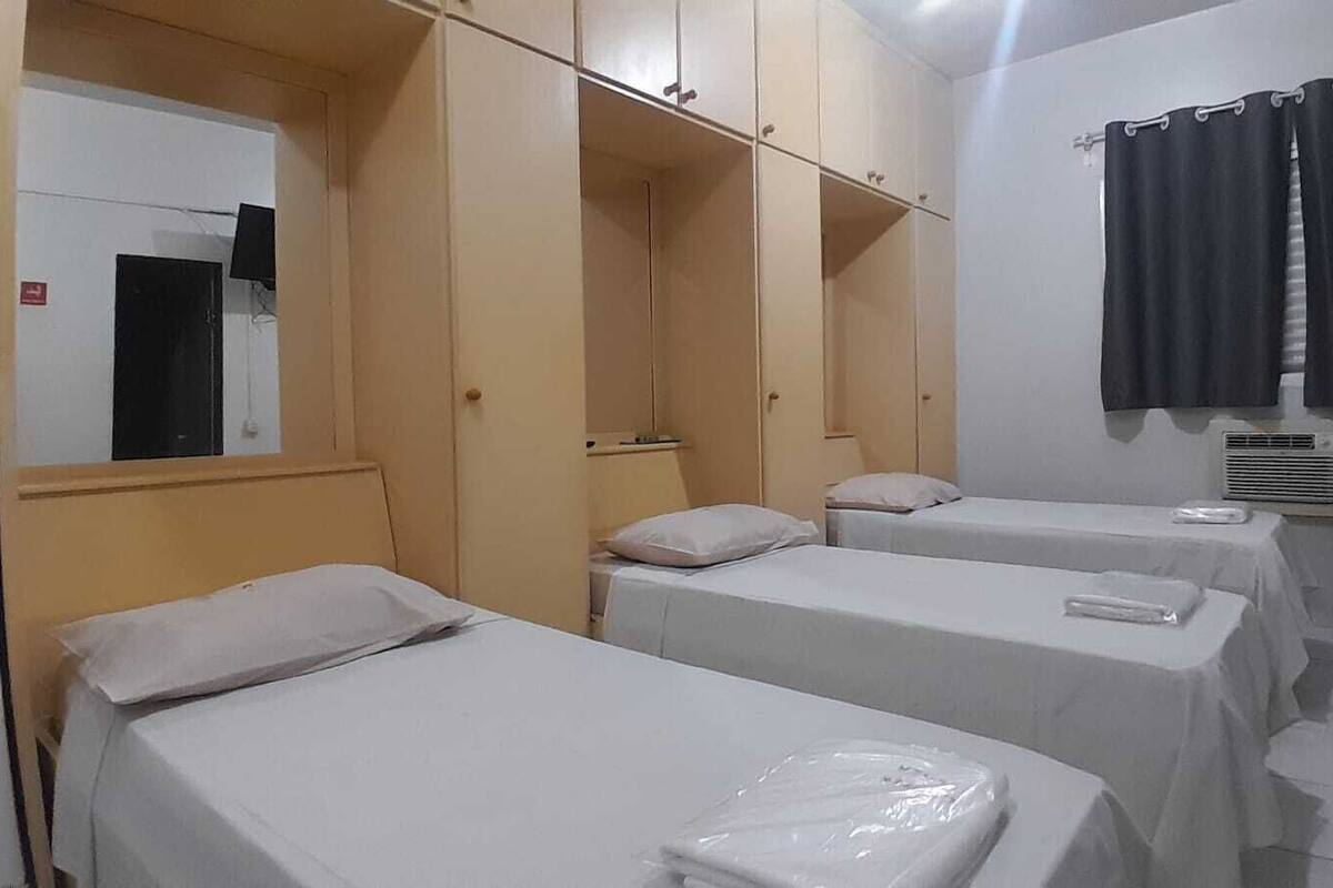 Habitación de hotel con tres camas individuales, un armario, un espejo, una ventana con cortinas y una mesita.