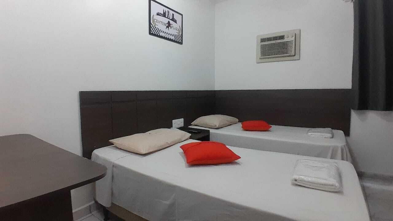 Une chambre d’hôtel avec deux lits simples, chacun équipé d’un oreiller et d’une couverture. On y trouve également une petite table et un climatiseur fixé au mur.
