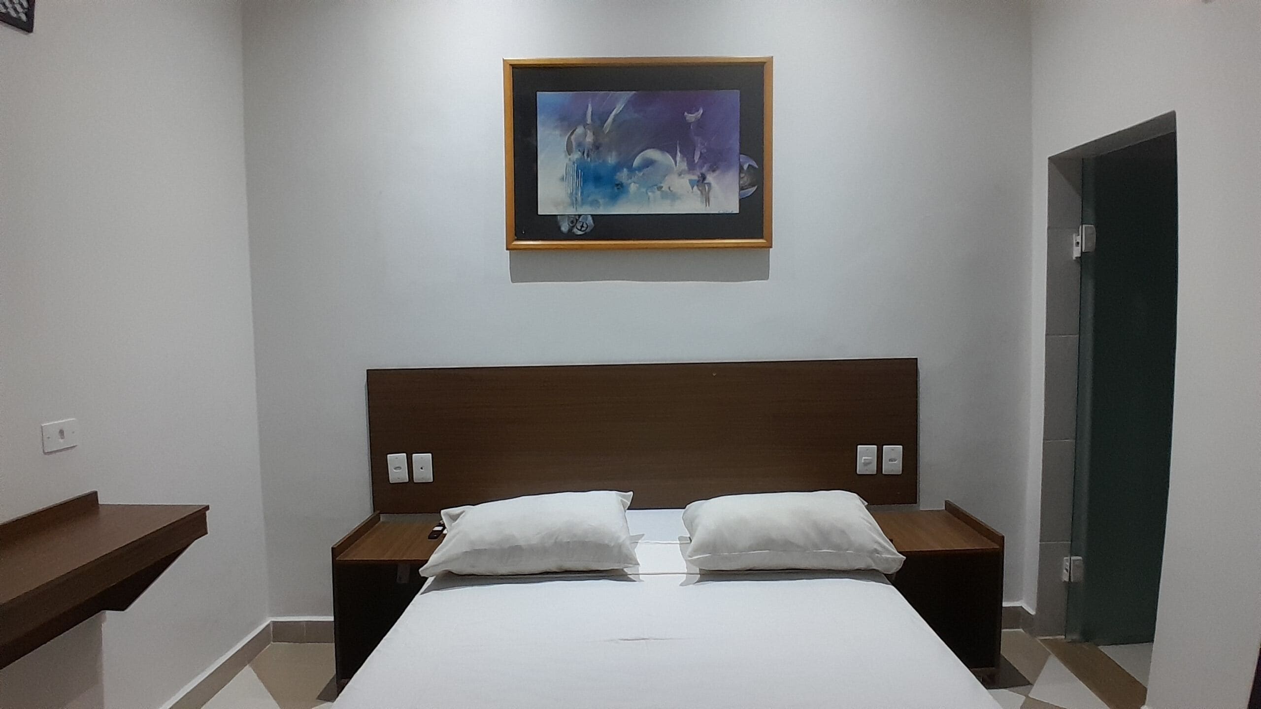 Ein Hotelzimmer mit einem Bett, zwei Nachttischen, einem Wandbehang über dem Bett und einer Tür auf der rechten Seite.