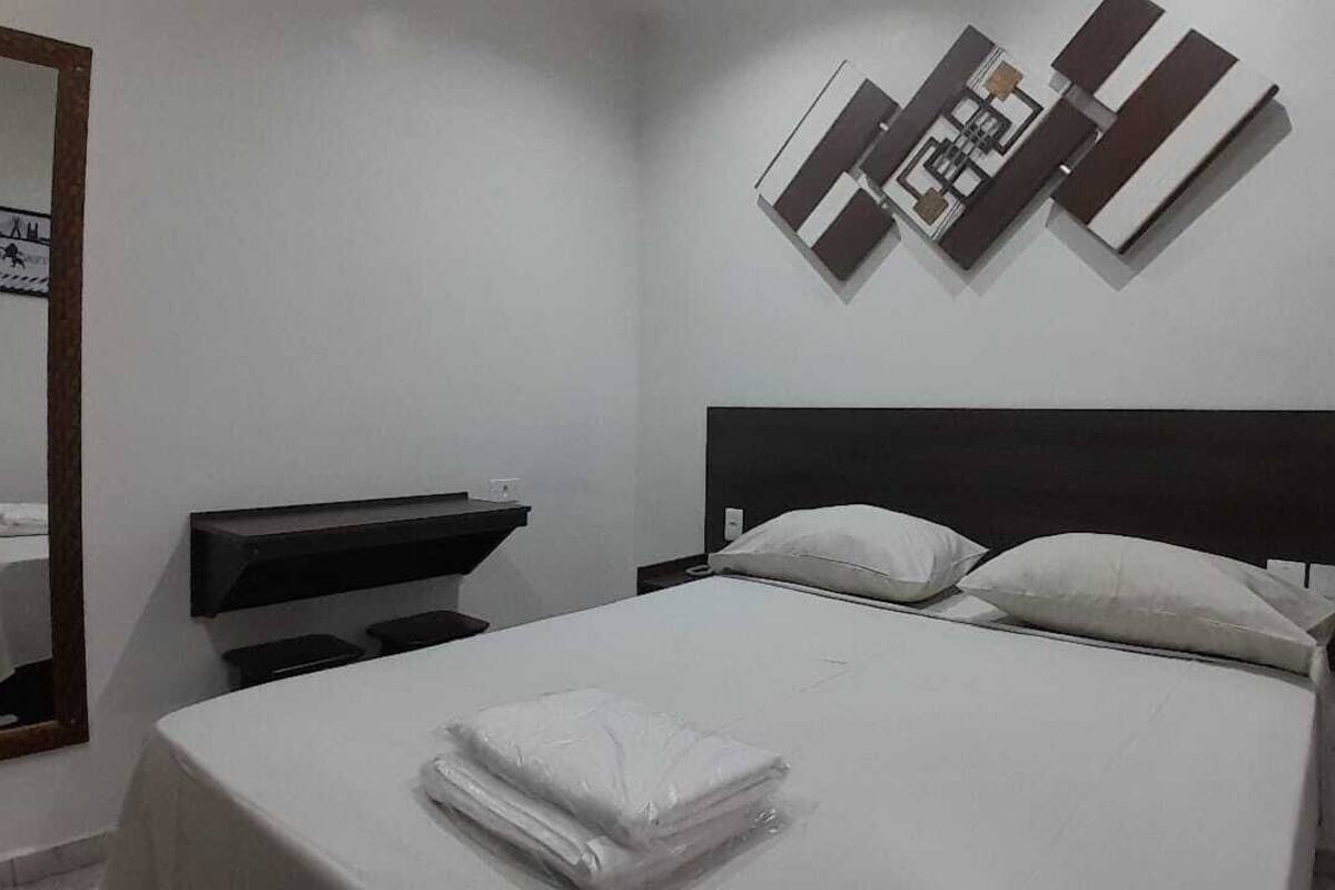 Una cama bien hecha con sábanas blancas, cabecera negra y dos almohadas. Una toalla blanca doblada está colocada al pie de la cama. Una mesita con superficie negra y cubierta blanca se encuentra a la izquierda de la cama. Se observa una obra de arte abstracto colgada en la pared sobre la cama.