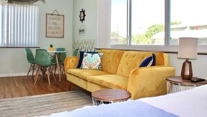 A room with a yellow sofa, a small round table, a green chair, a window with blinds, and a sign on the wall. - Seabreeze Suite Studio on Hollywood Beach (Hollywood)