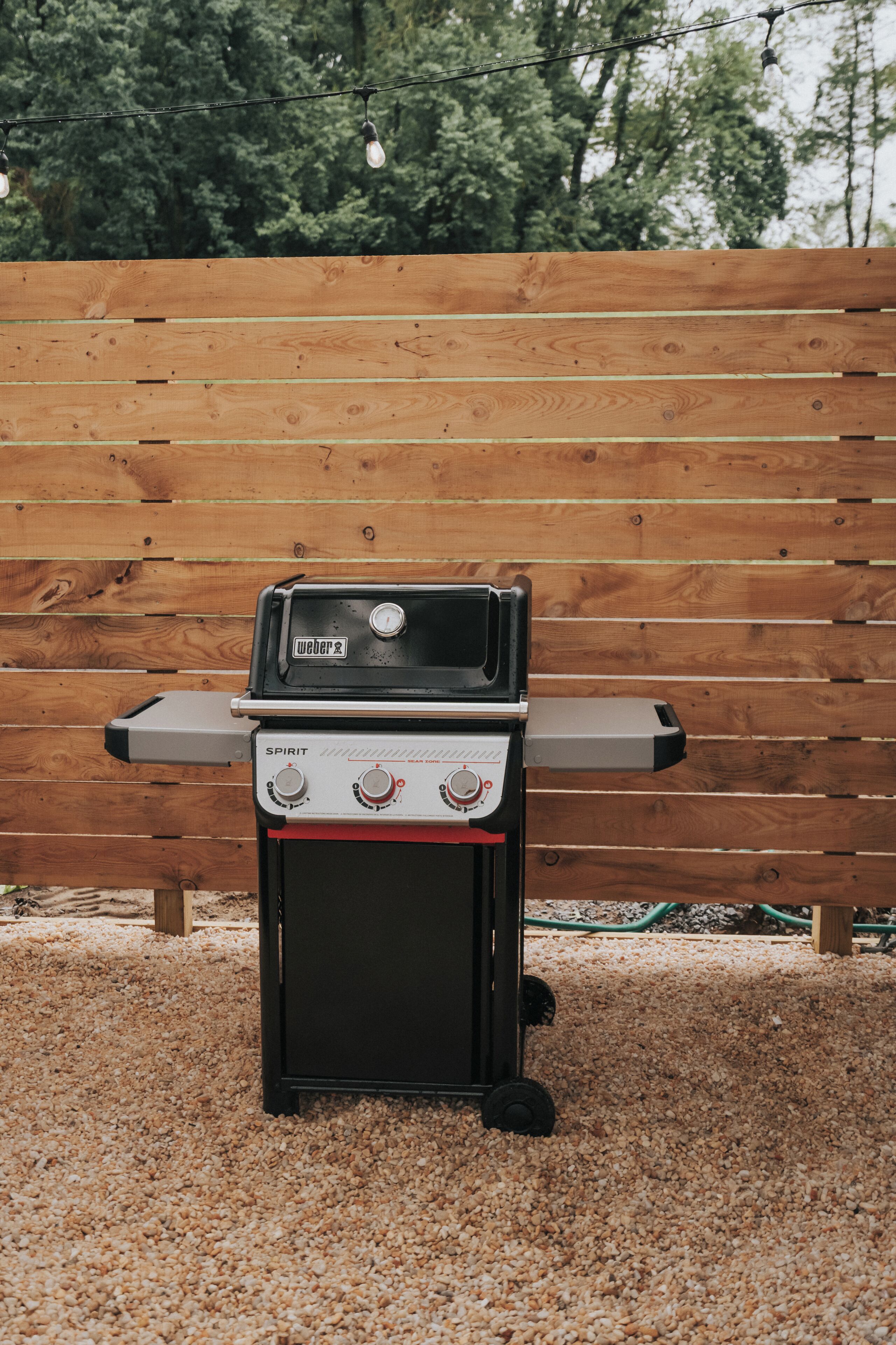 Un barbecue Weber Spirit portable doté d’un écran numérique, placé sur une surface de gravier, avec une clôture en bois en arrière-plan.