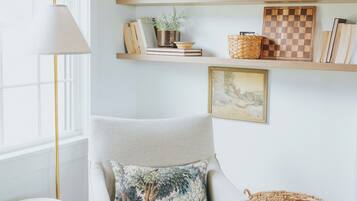 A cozy living room with a beige armchair, a small round table, and a wicker chair. Shelves above the armchair hold books, a framed picture, and decorative items.