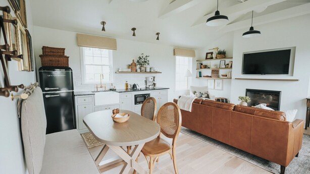 Una cocina moderna con armarios blancos, un comedor con mesa y sillas de madera, y un sofá de cuero marrón en la sala.