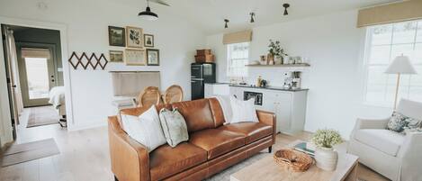 Un salón amplio con un sofá de cuero marrón, una mesa de centro de madera y un sillón blanco. Se ve un área de cocina al fondo con electrodomésticos modernos y una ventana.