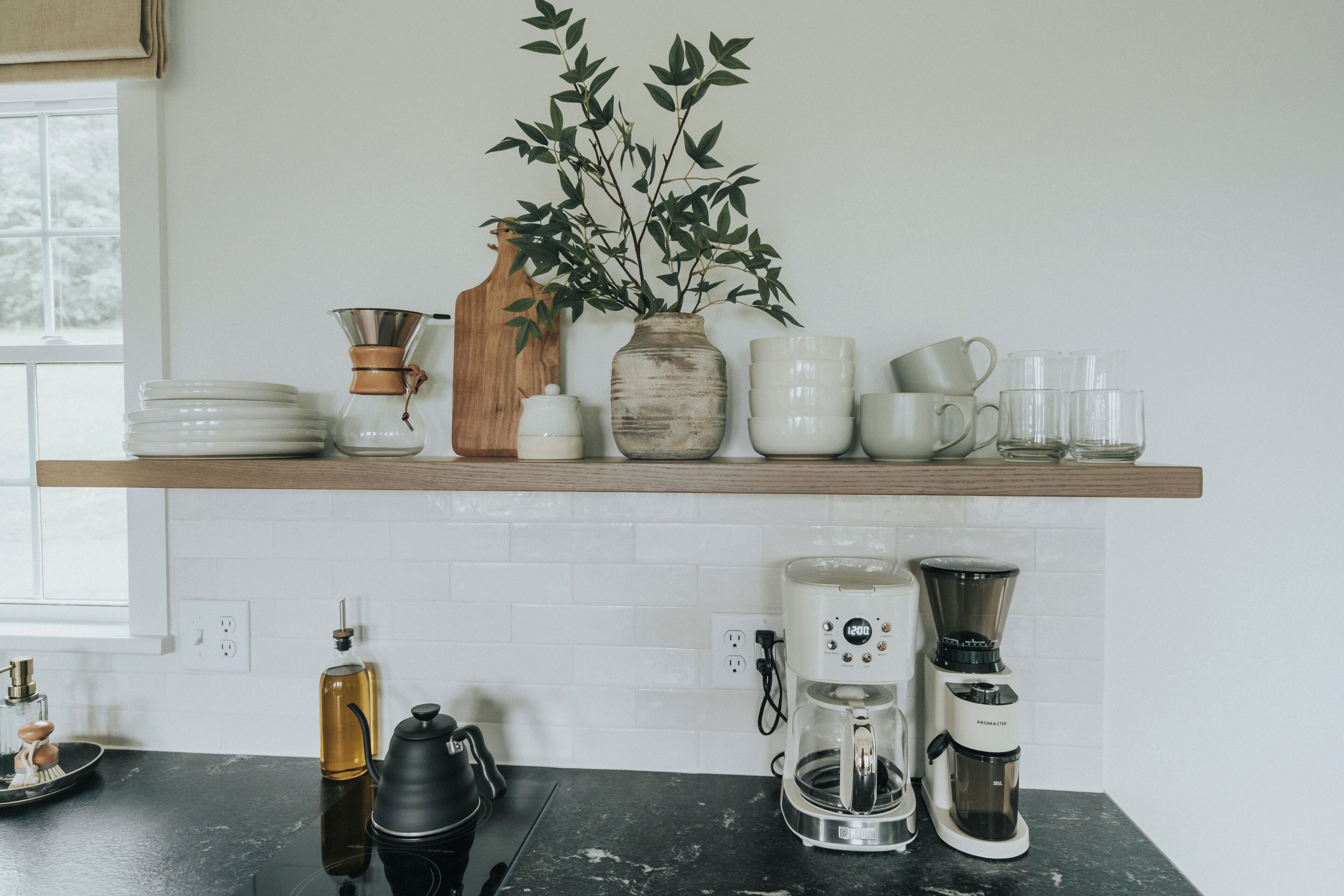 Een keuken met een houten plank waarop borden, bekers en een vaas met groene bladeren staan. Daaronder staan koffiezetapparaten en een waterkoker op het aanrecht.