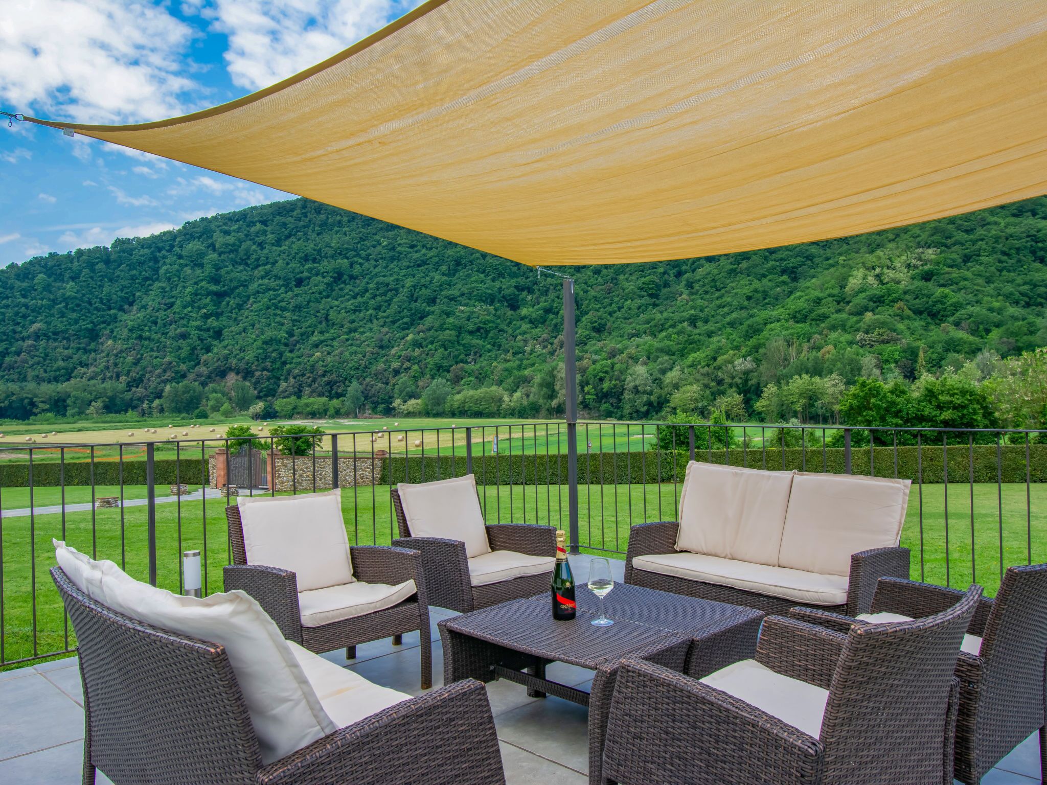 An outdoor patio with wicker furniture, a table with a bottle and glass, and a view of a green landscape with trees and a hill in the background.