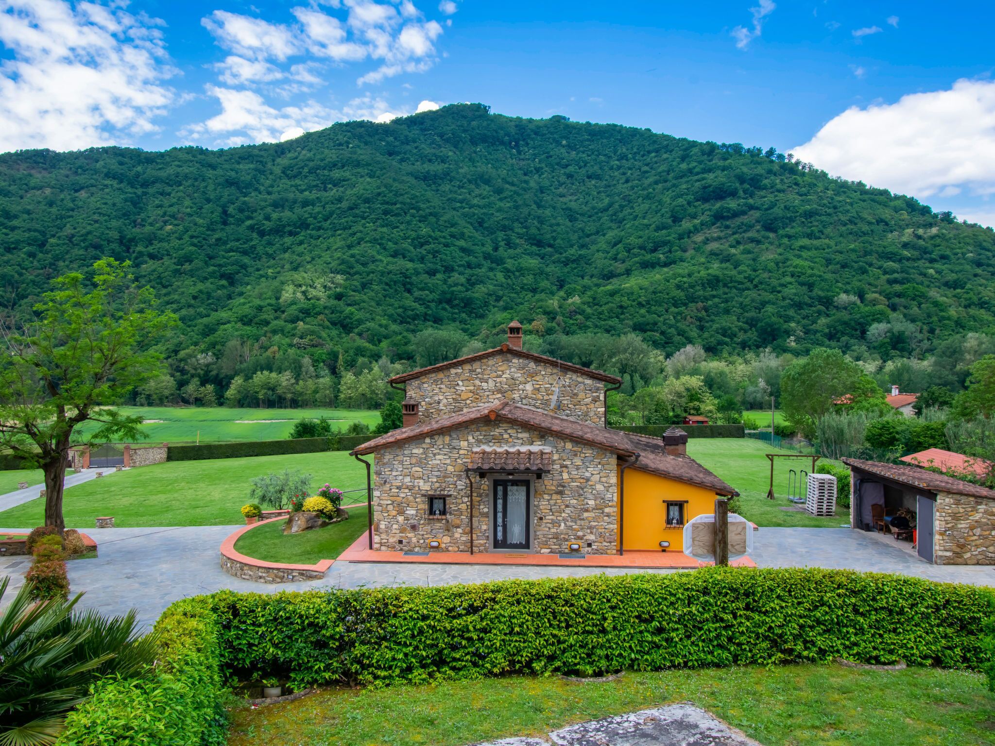 A stone house with a yellow facade, surrounded by a well-maintained garden and a mountain backdrop.