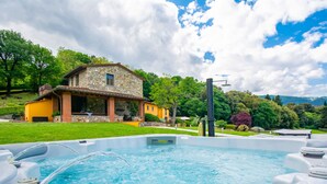 A swimming pool with hot tubs in the foreground, a stone house with a yellow facade in the background, and lush greenery surrounding the area.