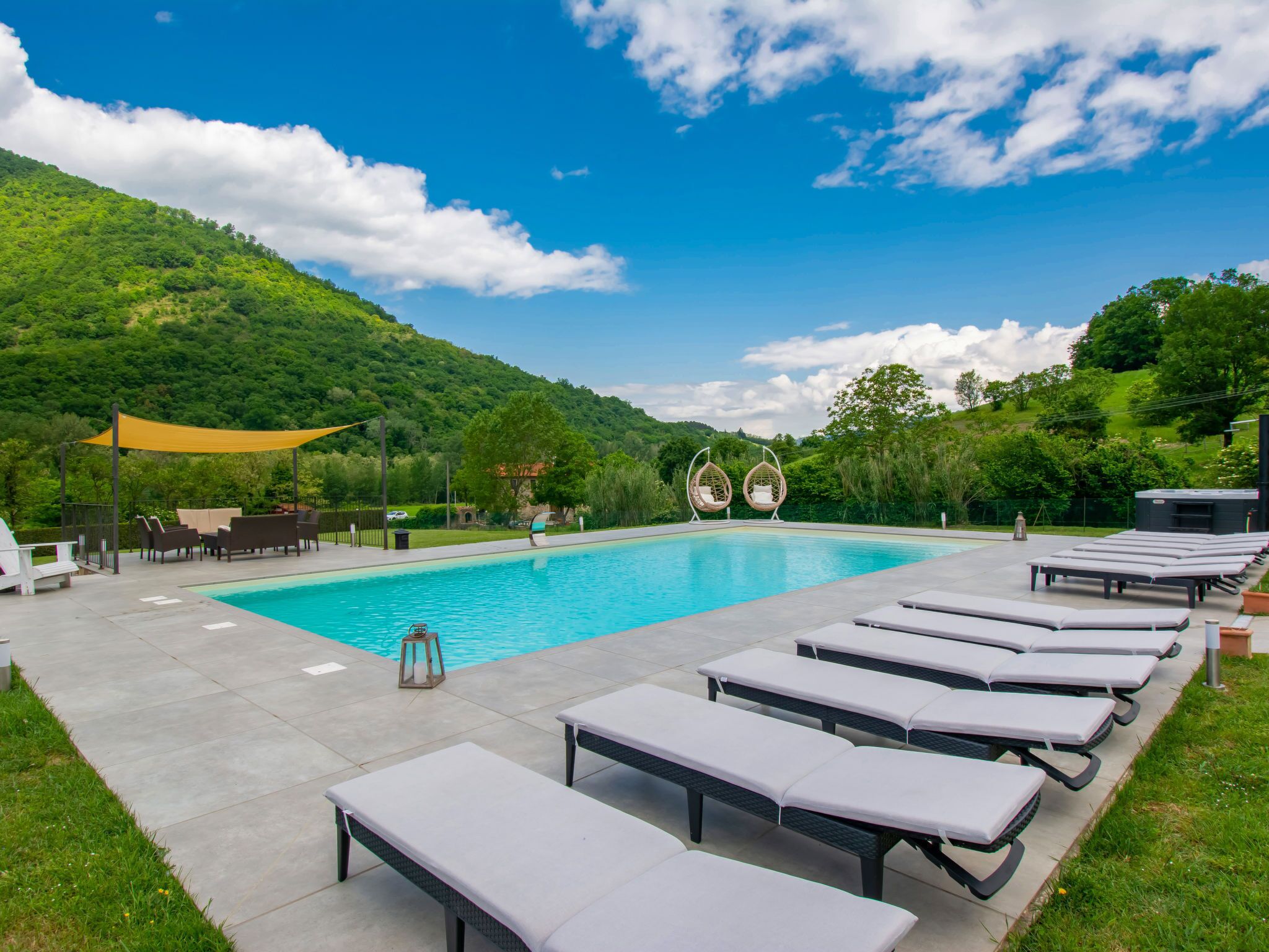 Ein Pool mit Liegestühlen und einem schattigen Bereich, umgeben von Grünflächen und Hügeln.